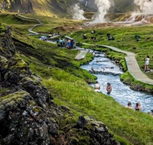 A scenic view of the Reykjadalur thermal river in Iceland, showing people soaking in the warm water surrounded by steaming green hills after a hike.