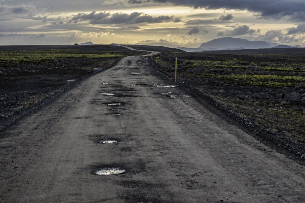Wet gravel road full of potholes in the Icelandic interior at dusk, with yellow marker posts along the edge