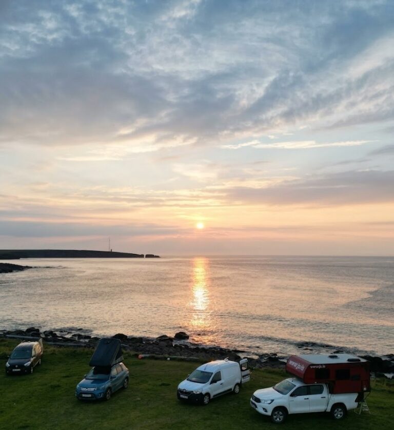 Four campervans and cars parked above a bay in north Iceland with the midnight sun low over the water